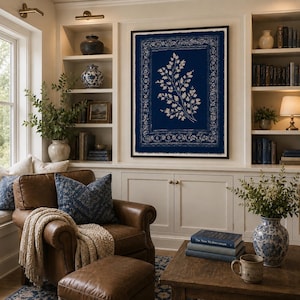 May include: A framed wall art piece featuring a dark blue background with a central cream-colored botanical fern illustration and a decorative border, displayed in a room with built-in shelving, a leather armchair, and a wooden coffee table.