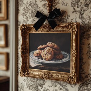 May include: A framed print of chocolate chip cookies on a blue and white plate, displayed in a gold ornate frame. A black velvet bow is tied above the frame, adding a decorative touch. The image is set against a floral wallpaper.