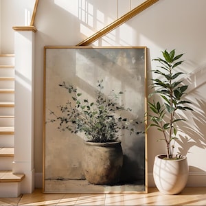 May include: A framed artwork featuring a large, textured pot filled with leafy green branches. The artwork is in a gold frame and is set against a neutral-toned wall. A potted plant with green leaves sits to the right.
