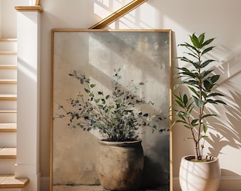 May include: A framed artwork featuring a large, textured pot filled with leafy green branches. The artwork is in a gold frame and is set against a neutral-toned wall. A potted plant with green leaves sits to the right.