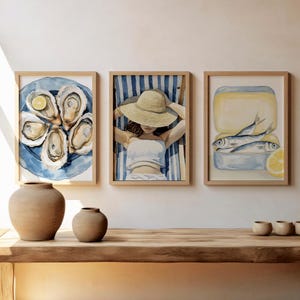 May include: Three framed watercolor paintings depicting a plate of oysters, a woman relaxing on a beach chair, and a can of sardines. The paintings are hung on a white wall above a wooden shelf with two brown ceramic vases.
