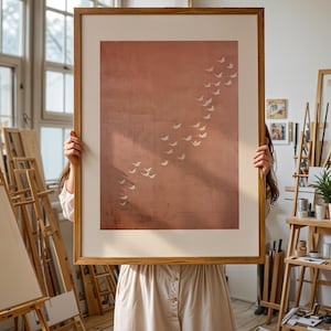 Puede incluir: Una impresión artística enmarcada que presenta una bandada de pájaros blancos en vuelo contra un fondo texturizado de color marrón rojizo. La obra de arte se muestra en un marco de madera natural, creando una estética minimalista. La impresión es sostenida por una persona con un vestido claro.