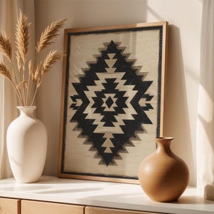 Op de afbeelding: Een ingelijste print met een zwart-wit geometrisch patroon. Het kunstwerk wordt getoond op een wit oppervlak, met een witte vaas met gedroogde planten en een bruine vaas. Het frame is gemaakt van lichtgekleurd hout.