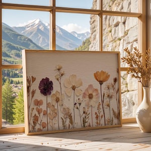 以下が含まれることがあります： 紫、クリーム、ピーチなど、さまざまな色のドライフラワーをあしらった額入りのアートワーク。アートワークは、山の景色を望む窓の近くの木製の表面に展示されています。フレームは明るい色の木材で作られています。