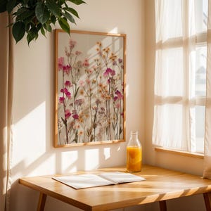 Może przedstawiać: Oprawiony botaniczny nadruk z prasowanymi kwiatami w odcieniach r&oacute;żu, ż&oacute;łci i bieli. Grafika wisi na kremowej ścianie, z drewnianym stołem i butelką soku pomarańczowego na pierwszym planie. Pomieszczenie jest oświetlone naturalnym światłem.