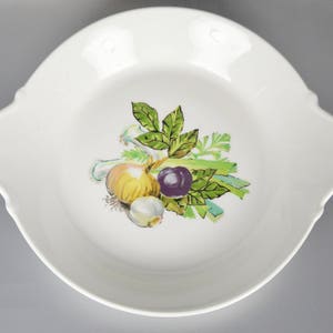 May include: A white ceramic serving dish with two side handles, featuring a central illustration of assorted vegetables including a yellow onion, a purple onion, a head of garlic, and green leafy herbs painted in vibrant colors on the basin.