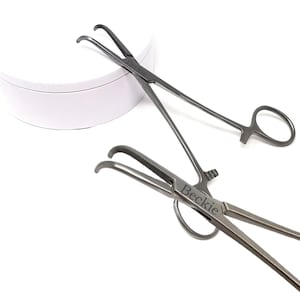 Puede incluir: Dos pinzas quirúrgicas grises con puntas curvas y asas de anillo se muestran sobre un fondo blanco. La palabra "Beckie" está grabada en las pinzas. Las pinzas están diseñadas para uso médico o científico.