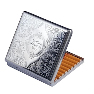 Puede incluir: Un pitillera plateada con un diseño floral ornamentado y el texto "Lydia's Once a Day". La pitillera está abierta, revelando cigarrillos. La pitillera tiene forma rectangular.
