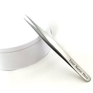 May include: A pair of silver tweezers with pointed tips, angled slightly to the right. The tweezers have the text "Your Name Here" engraved on the side. The tweezers are positioned in front of a white, round container.