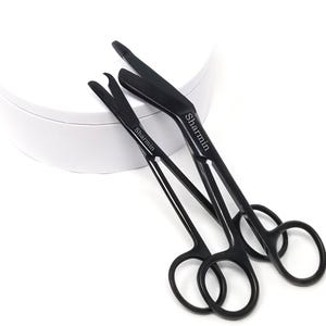 Peut inclure: Deux ciseaux chirurgicaux noirs avec le mot "Sharmin" gravé sur chacun. Les ciseaux sont sur une surface blanche.