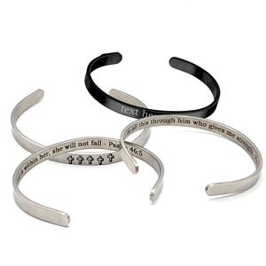 Può includere: Quattro braccialetti a polsino aperti in argento e nero. Un braccialetto nero ha il testo "text here". Due braccialetti argentati hanno testo inciso e simboli a croce. Il quarto braccialetto argentato ha testo inciso. Questi braccialetti sono un accessorio religioso.