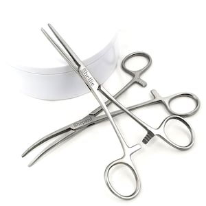 Puede incluir: Tres pinzas quirúrgicas de acero inoxidable con mangos de anillo y mandíbulas dentadas. Una pinza está etiquetada "Shellie" y otra "Shepard". Los instrumentos están dispuestos sobre un fondo blanco.