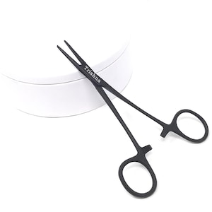 Puede incluir: Pinzas médicas de acero inoxidable negro con un mecanismo de bloqueo. Las pinzas están sobre una superficie blanca y el nombre de la marca "Trishana" está impreso en las pinzas.