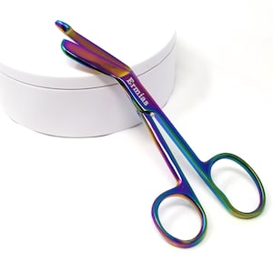 Pode incluir: Tesoura iridescente com a palavra "Ermias" impressa na lâmina. A tesoura tem um acabamento arco-íris com tons de azul, verde e roxo. As alças têm orifícios para os dedos.