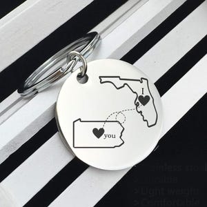 May include: A silver keychain with a round pendant featuring state outlines of Florida and Pennsylvania, connected by a dotted line. The keychain has the words "I" and "you" with heart symbols, symbolizing a long-distance relationship.