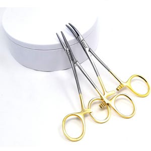 May include: Two surgical hemostatic forceps with gold-coloured handles and silver-coloured tips. The text "Name" and "Your" are engraved on the forceps. The forceps are on a white background.