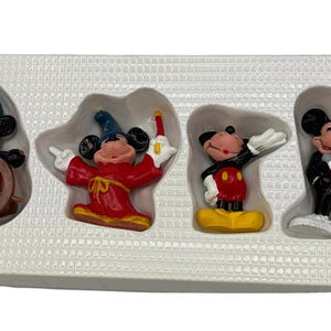 May include: A set of four Mickey Mouse figurines in a white plastic tray. The figurines depict Mickey Mouse in various outfits, including a sailor, a sorcerer, and a tuxedo. The figurines are colorful and detailed.