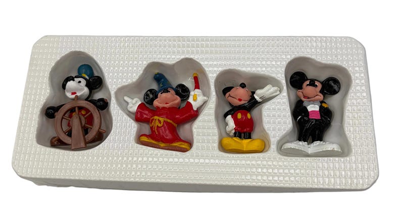 May include: A set of four Mickey Mouse figurines in a white plastic tray. The figurines depict Mickey Mouse in various outfits, including a sailor, a sorcerer, and a tuxedo. The figurines are colorful and detailed.
