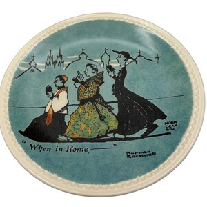 May include: A decorative plate with a teal center and a cream-colored rim. The plate features a cartoon illustration of three figures in religious attire walking, with the text "When in Rome" and artist signatures.