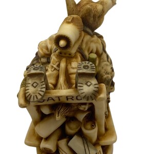 K&ouml;nnte beinhalten: Eine kleine, beige Keramikfigur eines Comic-Tieres, das einen Rucksack und Stiefel tr&auml;gt. Die Figur sitzt auf einem Stapel von Gegenst&auml;nden, darunter ein Stiefel, eine Flasche und ein Buch. Der Text "PATROL" ist auf der Basis der Figur sichtbar.