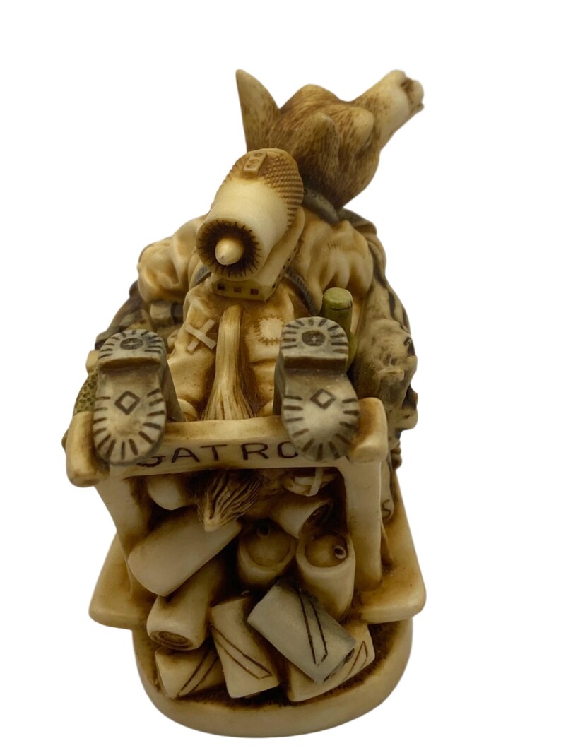 K&ouml;nnte beinhalten: Eine kleine, beige Keramikfigur eines Comic-Tieres, das einen Rucksack und Stiefel tr&auml;gt. Die Figur sitzt auf einem Stapel von Gegenst&auml;nden, darunter ein Stiefel, eine Flasche und ein Buch. Der Text "PATROL" ist auf der Basis der Figur sichtbar.