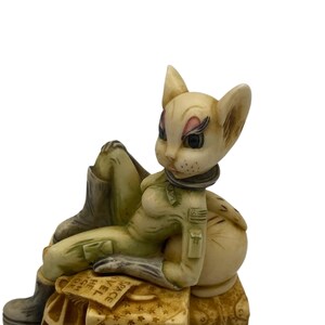 K&ouml;nnte beinhalten: Eine wei&szlig;e Katzenfigur, die einen gr&uuml;nen Fluganzug und Stiefel tr&auml;gt, sitzt auf Gep&auml;ck mit dem Wort "SPACE" auf einem Etikett. Die Katze hat einen Aufn&auml;her auf ihrem Anzug mit einem Stern und den Buchstaben "USA".