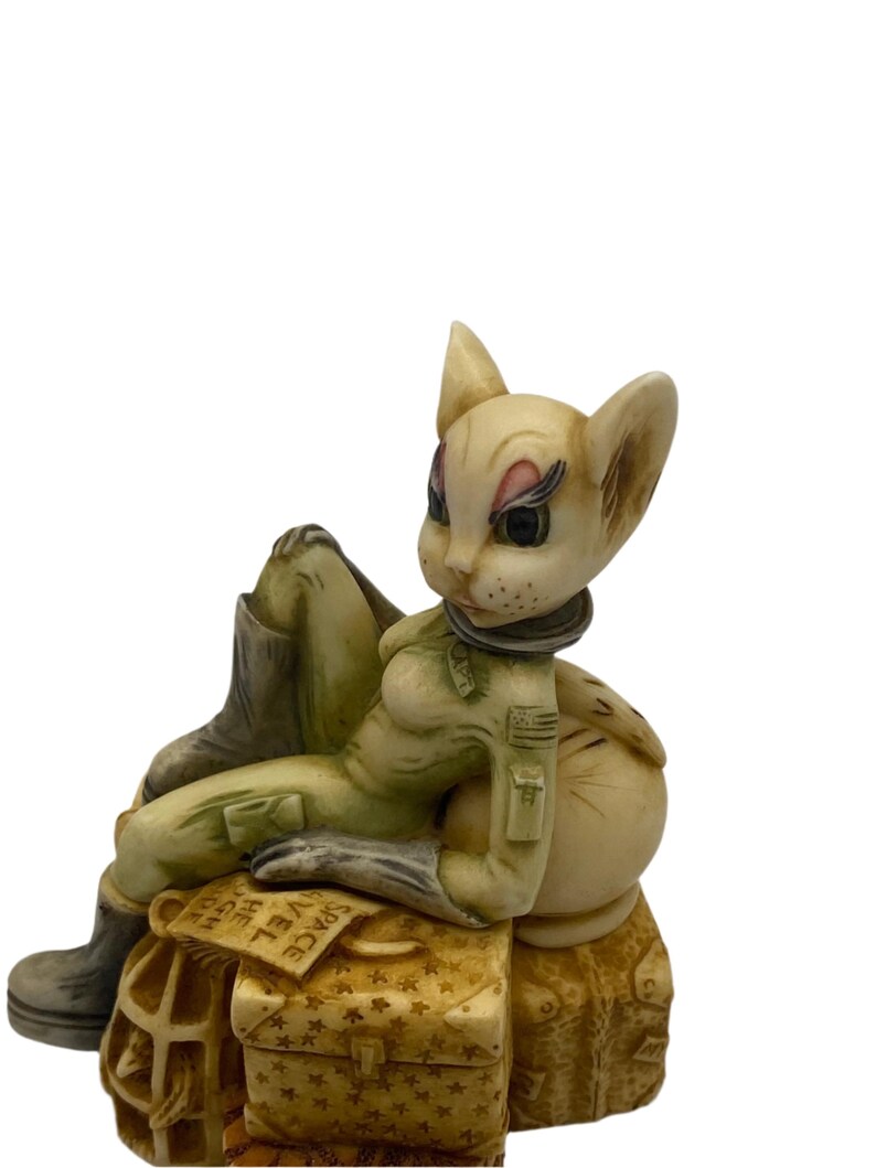 K&ouml;nnte beinhalten: Eine wei&szlig;e Katzenfigur, die einen gr&uuml;nen Fluganzug und Stiefel tr&auml;gt, sitzt auf Gep&auml;ck mit dem Wort "SPACE" auf einem Etikett. Die Katze hat einen Aufn&auml;her auf ihrem Anzug mit einem Stern und den Buchstaben "USA".