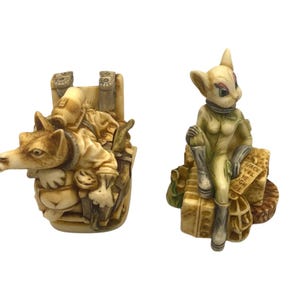 K&ouml;nnte beinhalten: Zwei Keramikfiguren, eine davon ein wolfartiges Wesen, das eine menschliche Figur auf dem R&uuml;cken tr&auml;gt, und die andere eine katzenartige Kreatur, die einen Raumanzug tr&auml;gt und auf einem Gep&auml;ckhaufen sitzt. Die katzenartige Kreatur hat den Text "Space Wel Come High De" auf ihrem Gep&auml;ck.