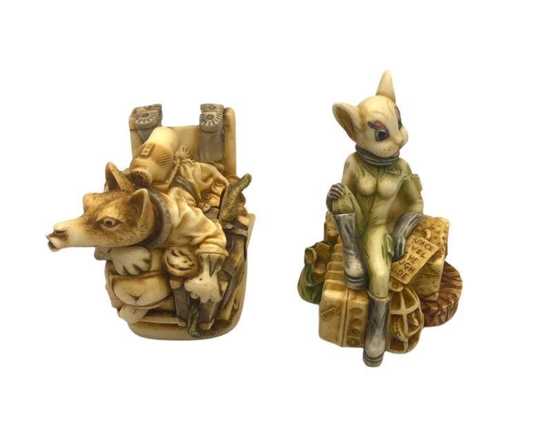 K&ouml;nnte beinhalten: Zwei Keramikfiguren, eine davon ein wolfartiges Wesen, das eine menschliche Figur auf dem R&uuml;cken tr&auml;gt, und die andere eine katzenartige Kreatur, die einen Raumanzug tr&auml;gt und auf einem Gep&auml;ckhaufen sitzt. Die katzenartige Kreatur hat den Text "Space Wel Come High De" auf ihrem Gep&auml;ck.