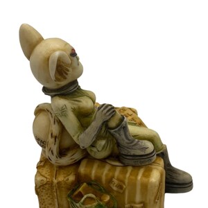K&ouml;nnte beinhalten: Eine Keramikfigur einer Frau mit Hasenohren, die eine gr&uuml;n-braune Uniform tr&auml;gt und auf Gep&auml;ck sitzt. Das Gep&auml;ck ist mit den Worten "Space", "Plum" und "Western Isles of Scotland" beschriftet.