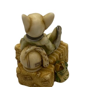K&ouml;nnte beinhalten: Eine kleine, cremefarbene Figur eines Katers, der ein gr&uuml;nes Hemd und einen Hut tr&auml;gt, sitzt auf einem Stapel Gep&auml;ck. Das Gep&auml;ck ist mit Reisestickern verziert und die Worte "Space Zone" sind auf dem Hut zu sehen.