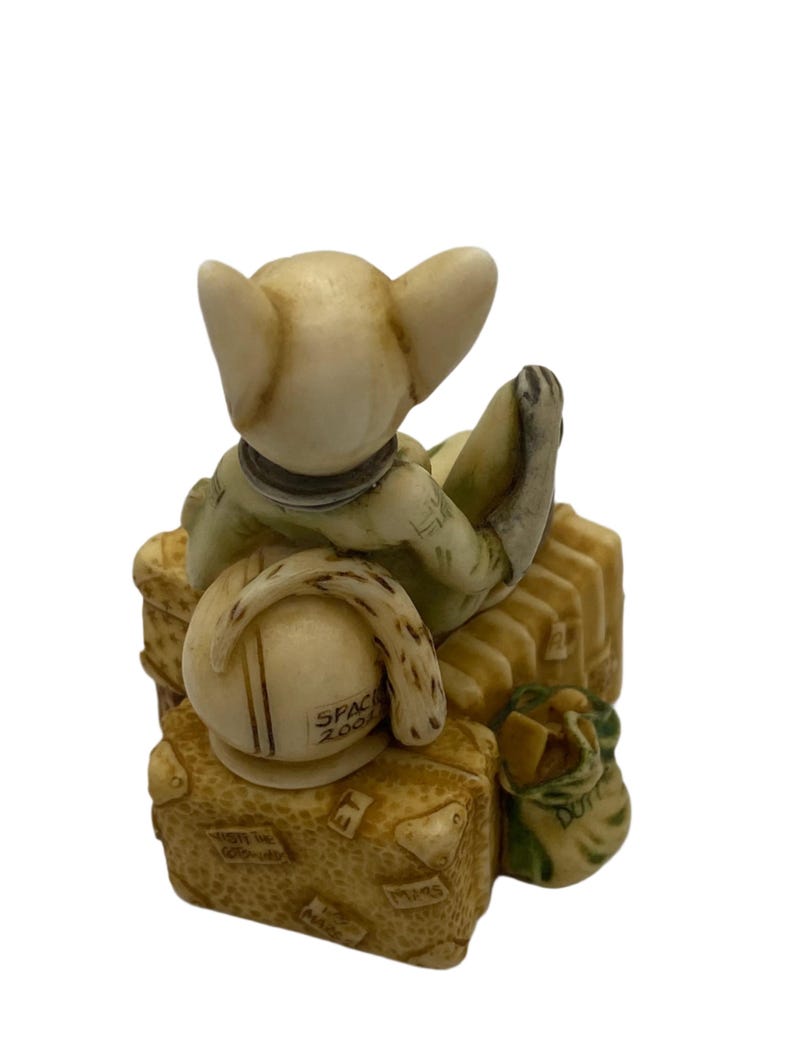 K&ouml;nnte beinhalten: Eine kleine, cremefarbene Figur eines Katers, der ein gr&uuml;nes Hemd und einen Hut tr&auml;gt, sitzt auf einem Stapel Gep&auml;ck. Das Gep&auml;ck ist mit Reisestickern verziert und die Worte "Space Zone" sind auf dem Hut zu sehen.