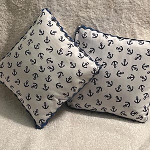 Puede incluir: Dos cojines cuadrados con fondo blanco y un estampado de anclas azul marino. Los cojines tienen un ribete festoneado azul marino.