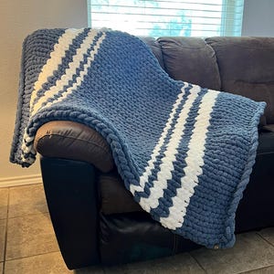 Könnte beinhalten: Eine blaue und weiße gestreifte, dicke Strickdecke, die über ein braunes Ledersofa gelegt ist. Die Decke ist aus dickem Garn gefertigt und hat eine weiche, kuschelige Textur.