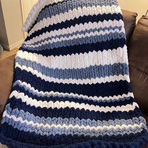 May include: A chunky knit blanket in shades of navy, white, and light blue. The blanket features horizontal stripes and a thick, textured appearance. The blanket is draped over a brown sofa, showcasing its cozy and decorative qualities.
