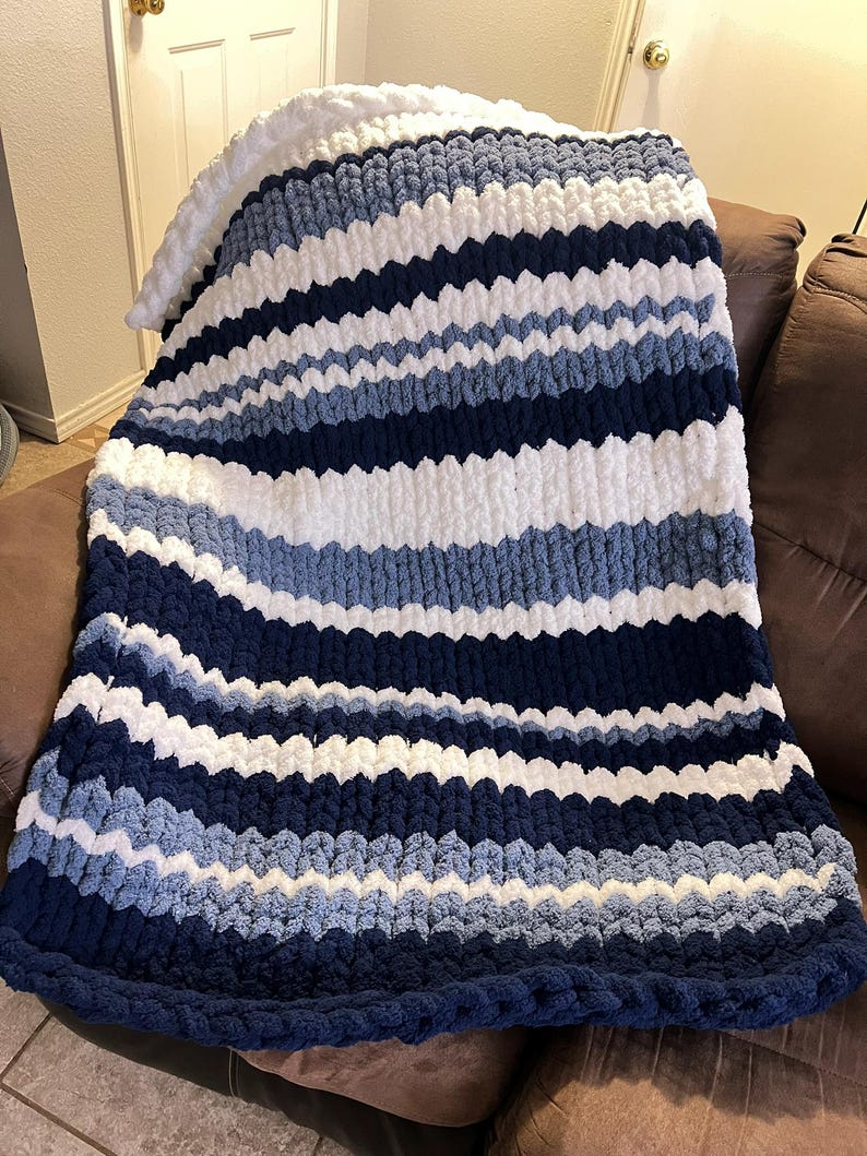 May include: A chunky knit blanket in shades of navy, white, and light blue. The blanket features horizontal stripes and a thick, textured appearance. The blanket is draped over a brown sofa, showcasing its cozy and decorative qualities.