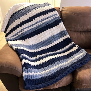 May include: A chunky knit blanket in shades of white, light blue, and navy blue, draped over a brown couch. The blanket features horizontal stripes and a thick, textured appearance, perfect for adding warmth and style to any living space.