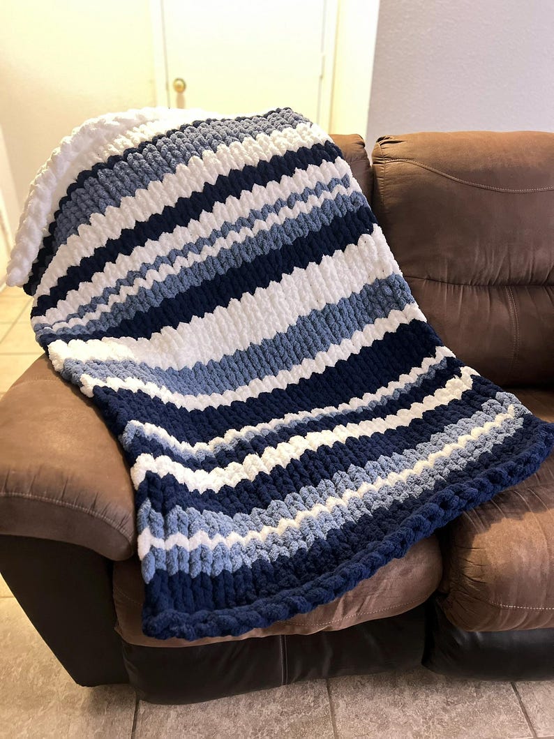 May include: A chunky knit blanket in shades of white, light blue, and navy blue, draped over a brown couch. The blanket features horizontal stripes and a thick, textured appearance, perfect for adding warmth and style to any living space.