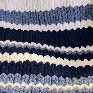 May include: A cozy, hand-knitted blanket with a textured, chunky knit design. The blanket features horizontal stripes in shades of white, light blue, and navy blue. The soft, plush material creates a visually appealing and tactile surface, perfect for adding warmth and style to any space.