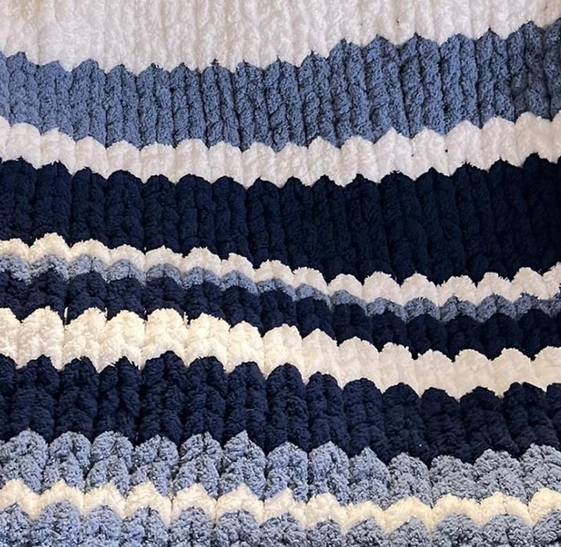 May include: A cozy, hand-knitted blanket with a textured, chunky knit design. The blanket features horizontal stripes in shades of white, light blue, and navy blue. The soft, plush material creates a visually appealing and tactile surface, perfect for adding warmth and style to any space.