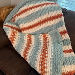 May include: A chunky knit throw blanket with stripes of white, light blue, and orange. The blanket is folded and draped over a brown leather couch.