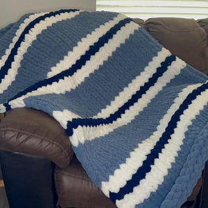 May include: A chunky knit blanket in shades of blue, white, and navy stripes. The soft, textured blanket is draped over a brown leather couch. The cozy throw blanket is perfect for snuggling.