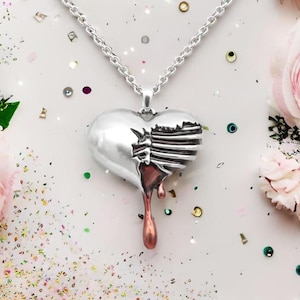 May include: A silver heart-shaped pendant necklace with a broken, hand-shaped design. The pendant is dripping with a red-brown substance, creating a dramatic and gothic aesthetic.