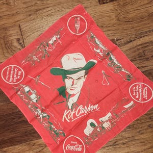 May include: Red square bandana featuring a portrait of Kit Carson in a cowboy hat, with the text "Kit Carson" and "Drink Coca-Cola." The design includes illustrations of historical scenes and text that reads "Hero of the Western Frontier."