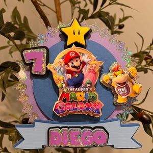 May include: A Super Mario Galaxy themed cake topper featuring Mario, Princess Peach, and Bowser. The topper includes a yellow star, the number 7, and the name "Diego" in pink and purple. The background is blue and purple with glitter accents.