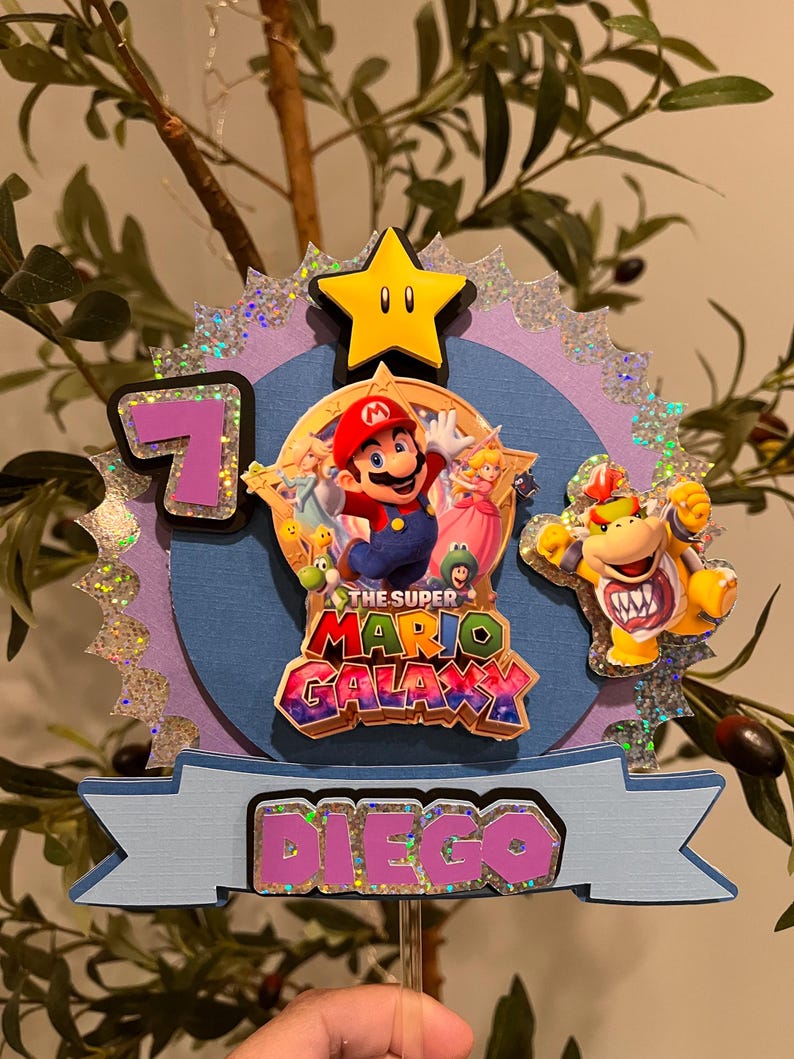 May include: A Super Mario Galaxy themed cake topper featuring Mario, Princess Peach, and Bowser. The topper includes a yellow star, the number 7, and the name "Diego" in pink and purple. The background is blue and purple with glitter accents.