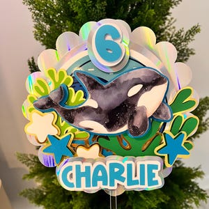 May include: A circular cake topper featuring a layered design with a holographic scalloped border. The center displays a watercolor-style orca, a blue number 6, and the name CHARLIE in blue letters. Accents include green seaweed and blue stars.