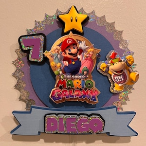 May include: A Super Mario Galaxy themed birthday cake topper. The topper features Mario, Princess Peach, and Bowser. The number 7 is displayed, along with the name "Diego". A yellow star and the words "The Super Mario Galaxy" are also included.