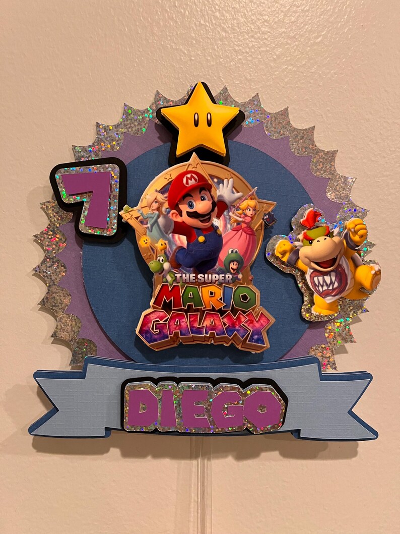May include: A Super Mario Galaxy themed birthday cake topper. The topper features Mario, Princess Peach, and Bowser. The number 7 is displayed, along with the name "Diego". A yellow star and the words "The Super Mario Galaxy" are also included.