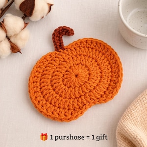 Puede incluir: Un posavasos de ganchillo con forma de calabaza en hilo naranja vibrante con un tallo marrón oscuro. El texto en la parte inferior dice: 🎁 1 purshase = 1 gift. El posavasos presenta un patrón de puntos circulares y texturizados.